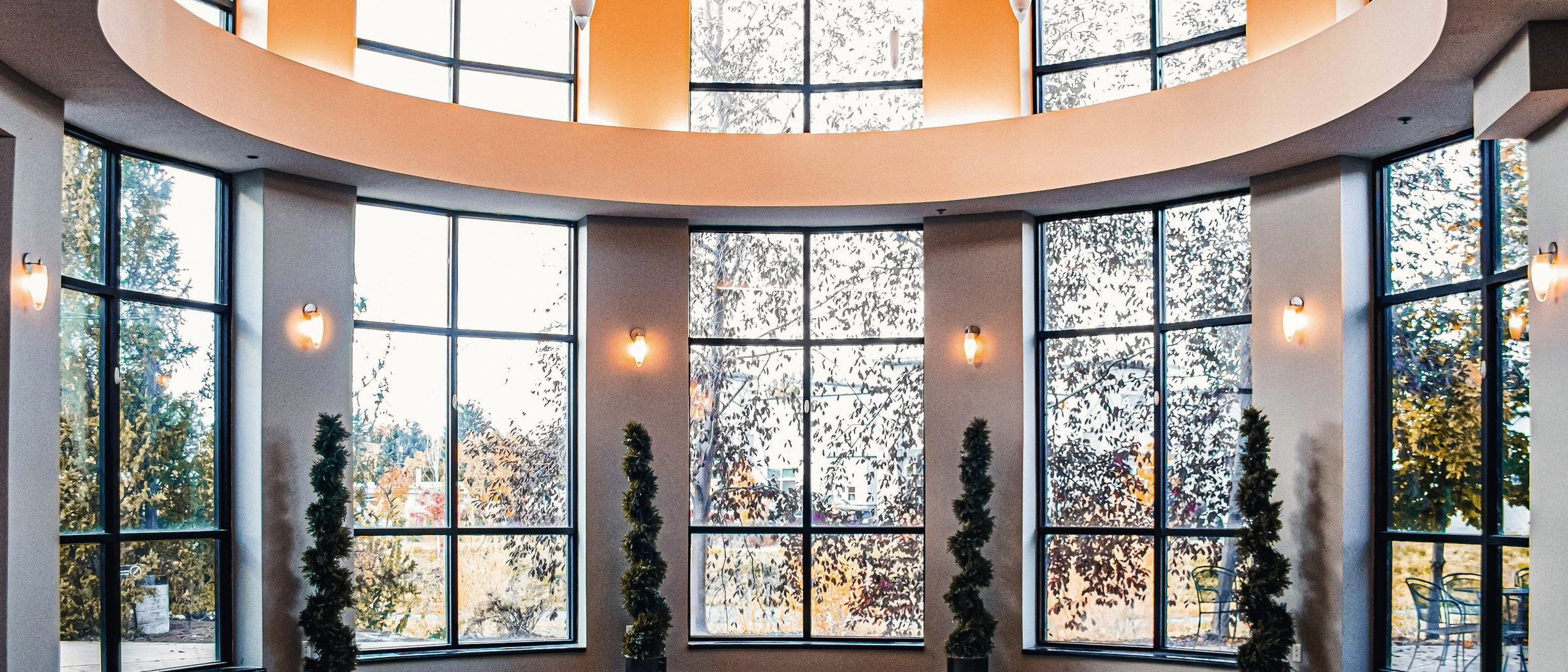 A curved room ringed with full-height windows under a circular opening in the ceiling, with pendant lights and topiary planters.