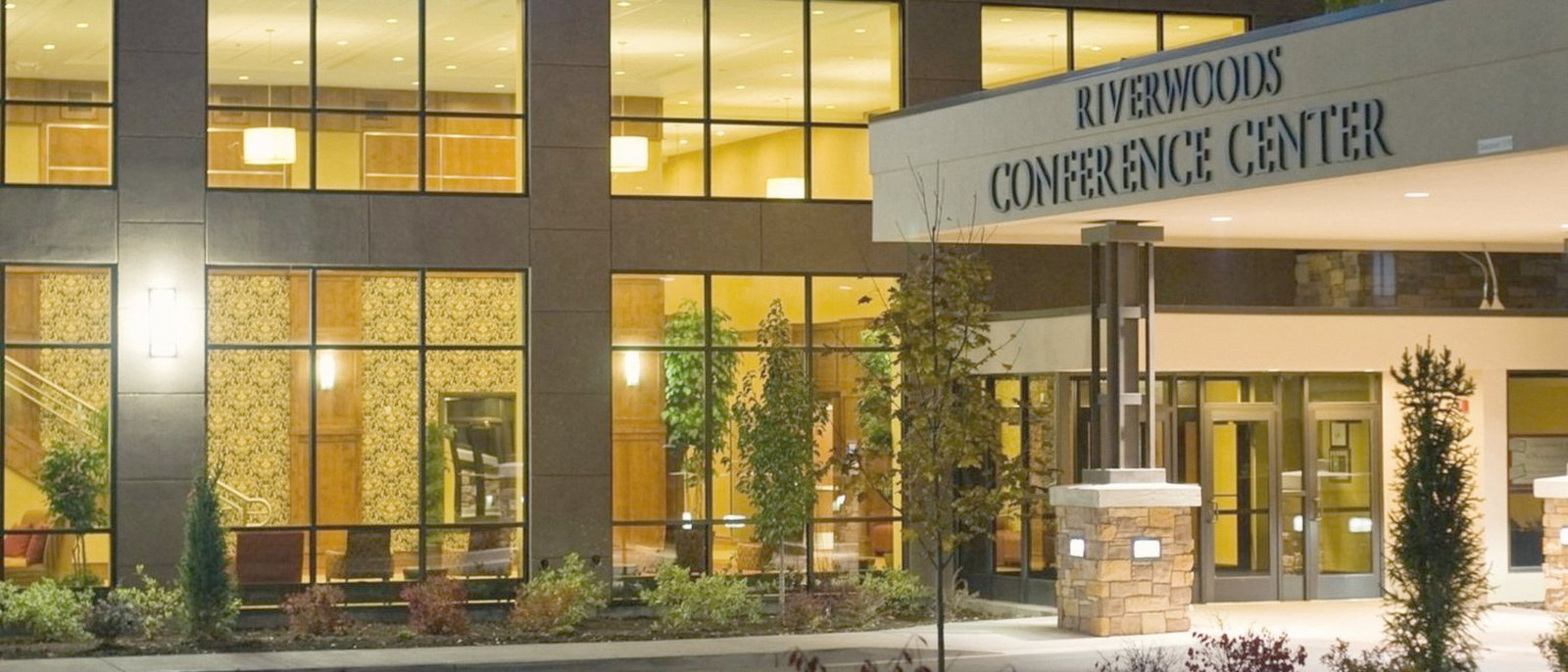 The Riverwoods Conference Center entrance at dusk, lit from within, with the name lettered over the canopy.